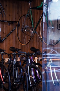 A collection of bicycles is neatly displayed against a wooden wall. Several bikes are aligned side by side on the floor, showcasing different frame colors like black and purple. A green bicycle is mounted on the wall, elevated above the rest. The setting appears to be a bicycle shop, with a glass panel reflecting some of the bikes.