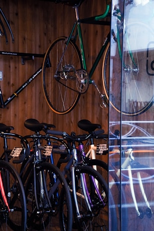 A collection of bicycles is neatly displayed against a wooden wall. Several bikes are aligned side by side on the floor, showcasing different frame colors like black and purple. A green bicycle is mounted on the wall, elevated above the rest. The setting appears to be a bicycle shop, with a glass panel reflecting some of the bikes.