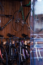 A collection of bicycles is neatly displayed against a wooden wall. Several bikes are aligned side by side on the floor, showcasing different frame colors like black and purple. A green bicycle is mounted on the wall, elevated above the rest. The setting appears to be a bicycle shop, with a glass panel reflecting some of the bikes.