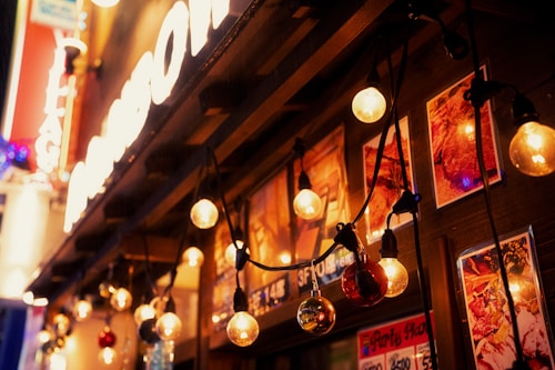A warmly lit storefront adorned with hanging string lights. The scene features an array of glowing bulbs, casting a cozy ambiance. Behind the lights are vivid posters, likely advertising food or drinks, adding a welcoming and lively feel.