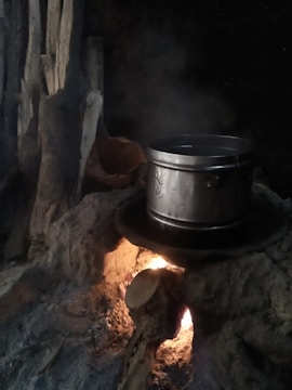 A metal pot sits on a rustic, makeshift stove made from rocks, with flames and smoke visible underneath. Wooden logs are stacked beside the stove, contributing to the fire.