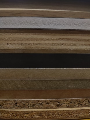 A stack of various wooden layers with textures ranging from smooth polished surfaces to rough, coarse edges. The layers exhibit varying shades of brown and beige, showcasing different types of wood.