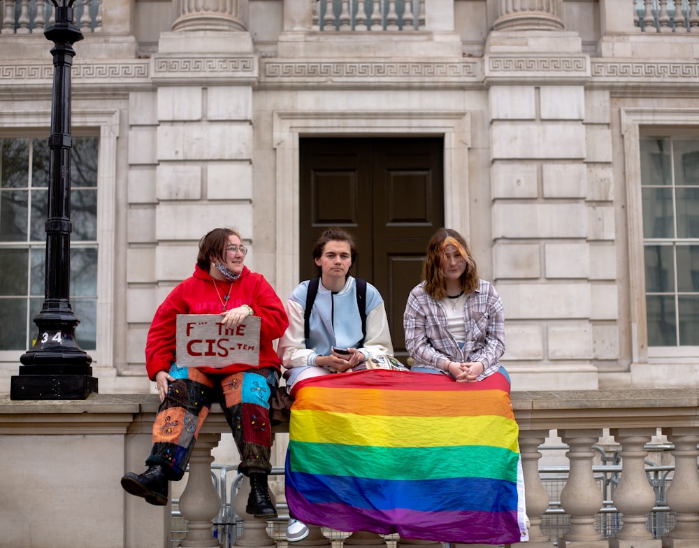 three people sitting on a ledge with a rainbow flag