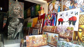 A collection of diverse paintings is displayed in a shop. The artwork includes a variety of styles and subjects such as a serene Buddha face, an abstract colorful lion, and scenes of traditional cultural activities. There are multiple paintings stacked and hung, showcasing vibrant colors and intricate details.