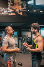 Two men are engaged in conversation at a gym. One man is older and wearing a grey t-shirt and glasses, while the other is younger, muscular, and wearing a sleeveless workout shirt with earbuds. They are standing near gym equipment, including a visible dumbbell on the floor. A large poster on the wall behind them features a muscular person with the text 'HARDEST WORKER IN THE ROOM'.