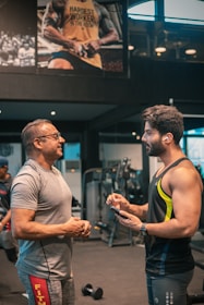 Two men are engaged in conversation at a gym. One man is older and wearing a grey t-shirt and glasses, while the other is younger, muscular, and wearing a sleeveless workout shirt with earbuds. They are standing near gym equipment, including a visible dumbbell on the floor. A large poster on the wall behind them features a muscular person with the text 'HARDEST WORKER IN THE ROOM'.