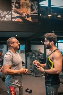 Two men are engaged in conversation at a gym. One man is older and wearing a grey t-shirt and glasses, while the other is younger, muscular, and wearing a sleeveless workout shirt with earbuds. They are standing near gym equipment, including a visible dumbbell on the floor. A large poster on the wall behind them features a muscular person with the text 'HARDEST WORKER IN THE ROOM'.