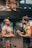 Two men are engaged in conversation at a gym. One man is older and wearing a grey t-shirt and glasses, while the other is younger, muscular, and wearing a sleeveless workout shirt with earbuds. They are standing near gym equipment, including a visible dumbbell on the floor. A large poster on the wall behind them features a muscular person with the text 'HARDEST WORKER IN THE ROOM'.
