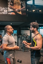 Two men are engaged in conversation at a gym. One man is older and wearing a grey t-shirt and glasses, while the other is younger, muscular, and wearing a sleeveless workout shirt with earbuds. They are standing near gym equipment, including a visible dumbbell on the floor. A large poster on the wall behind them features a muscular person with the text 'HARDEST WORKER IN THE ROOM'.