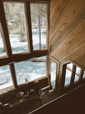 A peaceful snowy window view with gentle flakes falling outside, framed by warm wooden interiors and a stack of well-loved books.