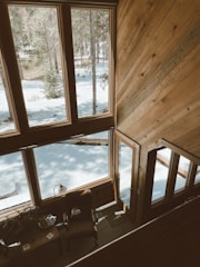 A cozy interior view from a high vantage point, showcasing large wooden-framed windows overlooking a snowy forest scene. Sunlight filters through the trees, casting shadows on the snow. Inside, there is a comfortable armchair with a small table nearby, suggesting a warm, inviting space for relaxation. The wood-paneled walls add to the rustic charm.