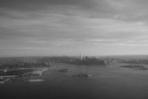 A grayscale aerial view of a large city skyline, featuring numerous skyscrapers and a wide body of water in the foreground. The Statue of Liberty is visible on a small island. The sky is overcast with a soft, cloudy texture.