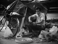 A man sitting on the sidewalk engaged in repairing tools, surrounded by various items and a bicycle with a covered load. The setup suggests a makeshift workstation with a collection of tools and materials scattered around.