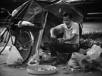 A man sitting on the sidewalk engaged in repairing tools, surrounded by various items and a bicycle with a covered load. The setup suggests a makeshift workstation with a collection of tools and materials scattered around.