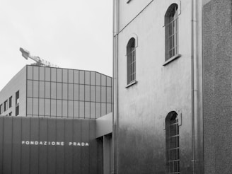 A modern building with a sleek, geometrical design, featuring large glass windows and minimalist architecture. The words 'Fondazione Prada' are visible on the facade. Adjacent, there is an older structure with arched windows, adding a contrast between contemporary and historical architectural styles. A construction crane is visible in the background, blending into the overcast sky.