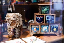 A detailed wooden elephant figurine, intricately carved and decorated with white patterns and colorful accents, stands next to a small, multicolored chest of drawers. Each drawer features unique geometric designs with vibrant colors, creating a lively and artistic display. The surface is wooden, and there appears to be a soft reflection, suggesting a glass display case.
