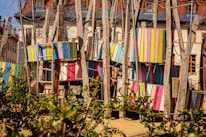 A colorful playground structure made of wooden beams and panels painted in various bright colors, including pink, blue, yellow, and green. Ladder-like structures and ropes are incorporated, suggesting a climbing area. The background shows residential buildings with steep roofs, and there is foliage in the foreground.