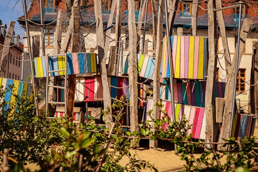 A colorful playground structure made of wooden beams and panels painted in various bright colors, including pink, blue, yellow, and green. Ladder-like structures and ropes are incorporated, suggesting a climbing area. The background shows residential buildings with steep roofs, and there is foliage in the foreground.