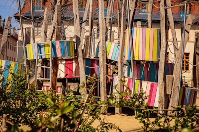 A colorful playground structure made of wooden beams and panels painted in various bright colors, including pink, blue, yellow, and green. Ladder-like structures and ropes are incorporated, suggesting a climbing area. The background shows residential buildings with steep roofs, and there is foliage in the foreground.
