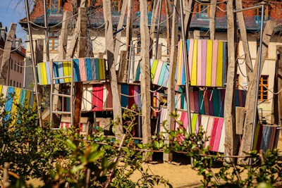 A colorful playground structure made of wooden beams and panels painted in various bright colors, including pink, blue, yellow, and green. Ladder-like structures and ropes are incorporated, suggesting a climbing area. The background shows residential buildings with steep roofs, and there is foliage in the foreground.