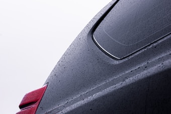 Raindrops cover the surface of a sleek, dark-colored car, highlighting its smooth curves and edges. The image captures a close-up view of the car's rear section, including part of a window and a rear light with a bright red tint. The water droplets give the surface a textured appearance.