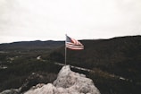 An American flag waves on a pole atop a rocky outcrop with a vast expanse of forested hills and mountains in the background, under a cloudy sky.