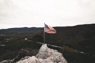 An American flag waves on a pole atop a rocky outcrop with a vast expanse of forested hills and mountains in the background, under a cloudy sky.