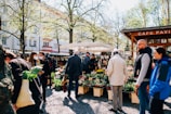 People are gathered at an outdoor market surrounded by trees and buildings. Several individuals are wearing jackets, and one person is holding a bag with greens. There are tables displaying various potted plants and flowers. The environment is bright and sunny, with people interacting and browsing the market stalls.