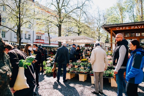 People are gathered at an outdoor market surrounded by trees and buildings. Several individuals are wearing jackets, and one person is holding a bag with greens. There are tables displaying various potted plants and flowers. The environment is bright and sunny, with people interacting and browsing the market stalls.
