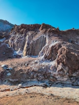 A rugged rocky landscape featuring various textures and tones. The brown and white minerals contrast against the layered rock formations, under a clear blue sky. The scene suggests geological activity or erosion, with a pathway or channel running through the foreground.