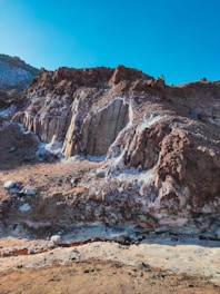 A rugged rocky landscape featuring various textures and tones. The brown and white minerals contrast against the layered rock formations, under a clear blue sky. The scene suggests geological activity or erosion, with a pathway or channel running through the foreground.