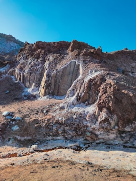 A rugged rocky landscape featuring various textures and tones. The brown and white minerals contrast against the layered rock formations, under a clear blue sky. The scene suggests geological activity or erosion, with a pathway or channel running through the foreground.