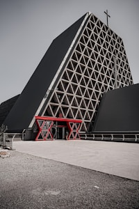 A modern triangular building with a large lattice structure on its front and a prominent cross at the top. The entrance is marked by a striking red geometric frame, contrasted against the building’s dark exterior.
