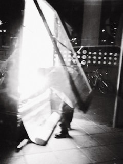 An abstract, black-and-white image with a motion blur effect. It depicts a figure bending down next to a bright light, possibly taking down or setting up a large sign or piece of equipment. In the background, there is a bicycle visible.