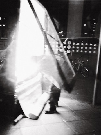 An abstract, black-and-white image with a motion blur effect. It depicts a figure bending down next to a bright light, possibly taking down or setting up a large sign or piece of equipment. In the background, there is a bicycle visible.