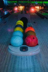 A row of colorful bowling balls is positioned on a bowling lane with a darkened background. The balls range in hues of blue, yellow, orange, and red. The setting appears to be a bowling alley with dim lighting and glowing neon lights.