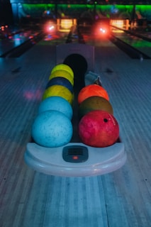 A row of colorful bowling balls is positioned on a bowling lane with a darkened background. The balls range in hues of blue, yellow, orange, and red. The setting appears to be a bowling alley with dim lighting and glowing neon lights.