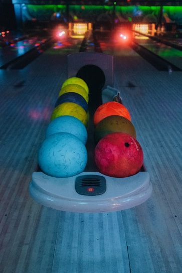 A row of colorful bowling balls is positioned on a bowling lane with a darkened background. The balls range in hues of blue, yellow, orange, and red. The setting appears to be a bowling alley with dim lighting and glowing neon lights.