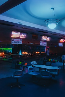 Inside a dimly lit bowling alley, featuring multiple bowling lanes illuminated by neon and colored lights. Overhead screens display scores and seating areas with bowling balls neatly arranged. The atmosphere has a moody, almost futuristic feel due to the ambient lighting.