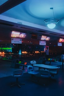 Inside a dimly lit bowling alley, featuring multiple bowling lanes illuminated by neon and colored lights. Overhead screens display scores and seating areas with bowling balls neatly arranged. The atmosphere has a moody, almost futuristic feel due to the ambient lighting.