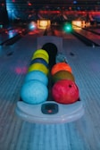 A set of colorful bowling balls is neatly arranged on a bowling ball rack, with lanes and pinsetters visible in the background. The scene is dimly lit with multi-colored lights creating a lively atmosphere.