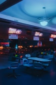 A dimly lit bowling alley with glowing neon lights illuminating the area. Bowling lanes extend toward the back, accompanied by a series of scoreboards hanging from the ceiling. Several colorful bowling balls are placed in racks beside chairs and tables designed for seating near the lanes.