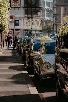 A row of parked taxis line the city street, with reflective surfaces gleaming under sunlight. Pedestrians are visible on the adjacent sidewalk, walking past tall buildings with glass facades. Street signs are mounted above, indicating taxi lanes and pedestrian crossings.