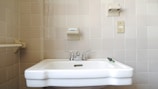 A bathroom sink with silver faucets mounted on a light-colored tiled wall. Above the sink, there is a soap holder with a bar of soap, and to the right, a light switch is visible. The overall setup conveys a minimal and functional design.