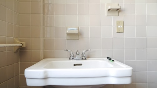 A bathroom sink with silver faucets mounted on a light-colored tiled wall. Above the sink, there is a soap holder with a bar of soap, and to the right, a light switch is visible. The overall setup conveys a minimal and functional design.