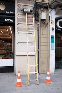 A tall yellow ladder is positioned against a building's wall where numerous cables are tangled and connected to electrical equipment. The area is marked off with several orange and white traffic cones on a tiled sidewalk. A storefront with visible products inside is partially in view to the left.
