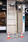 A tall yellow ladder is positioned against a building's wall where numerous cables are tangled and connected to electrical equipment. The area is marked off with several orange and white traffic cones on a tiled sidewalk. A storefront with visible products inside is partially in view to the left.