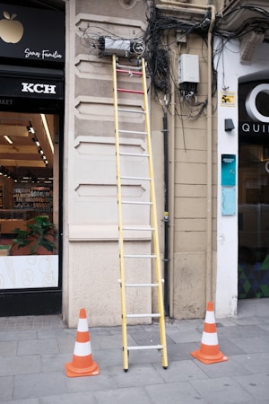 A tall yellow ladder is positioned against a building's wall where numerous cables are tangled and connected to electrical equipment. The area is marked off with several orange and white traffic cones on a tiled sidewalk. A storefront with visible products inside is partially in view to the left.