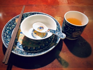 A beautifully arranged set of ceramic dishes comprising a large plate, a smaller bowl, and a soup spoon, all featuring intricate blue and white floral designs with touches of yellow and orange. A pair of wooden chopsticks rests on the bowl, and a matching cup filled with a warm amber liquid is placed nearby. The set is placed on a polished wooden surface, reflecting warm ambient light.