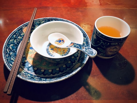 A beautifully arranged set of ceramic dishes comprising a large plate, a smaller bowl, and a soup spoon, all featuring intricate blue and white floral designs with touches of yellow and orange. A pair of wooden chopsticks rests on the bowl, and a matching cup filled with a warm amber liquid is placed nearby. The set is placed on a polished wooden surface, reflecting warm ambient light.